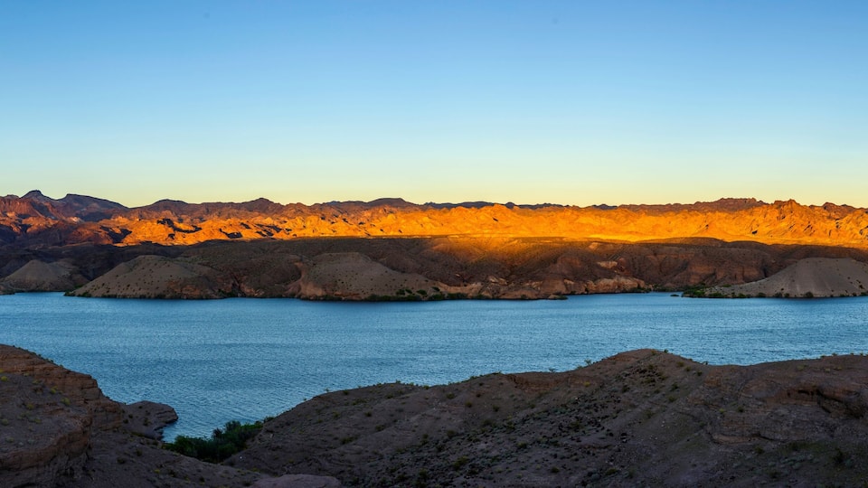 A mountainous landscape with a body of water in the foreground and a sunset casting warm tones on the hills.