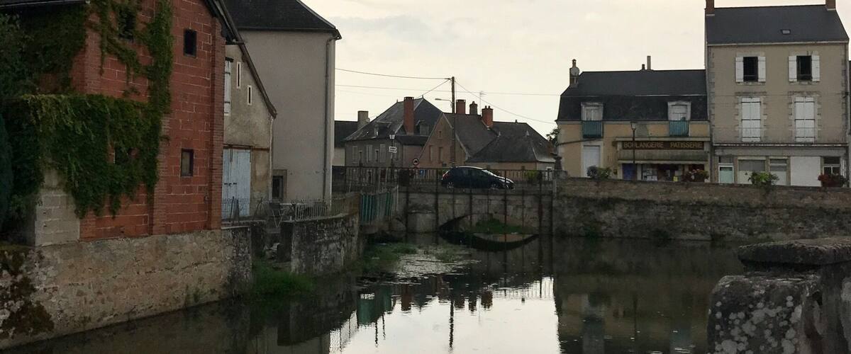 A calm river reflecting buildings, a stone bridge, and a cloudy sky.