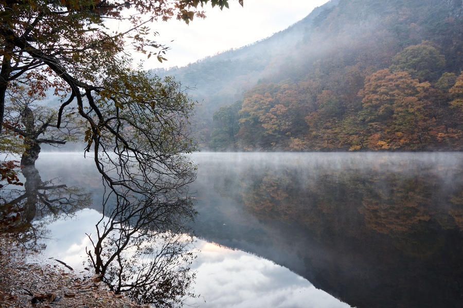 A calm lake surrounded by autumn-colored trees and misty mountains.