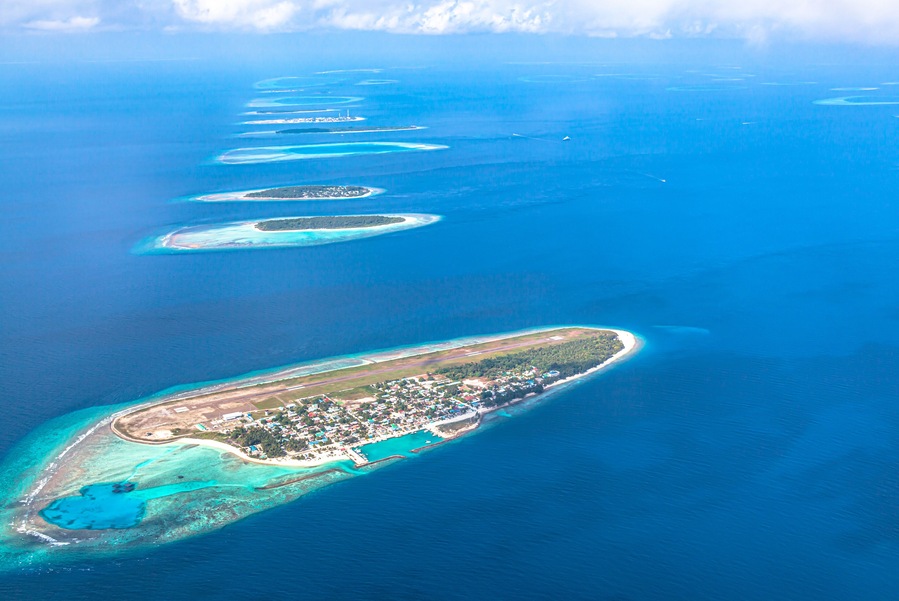 An aerial view of a chain of turquoise atolls with a developed island at the end.