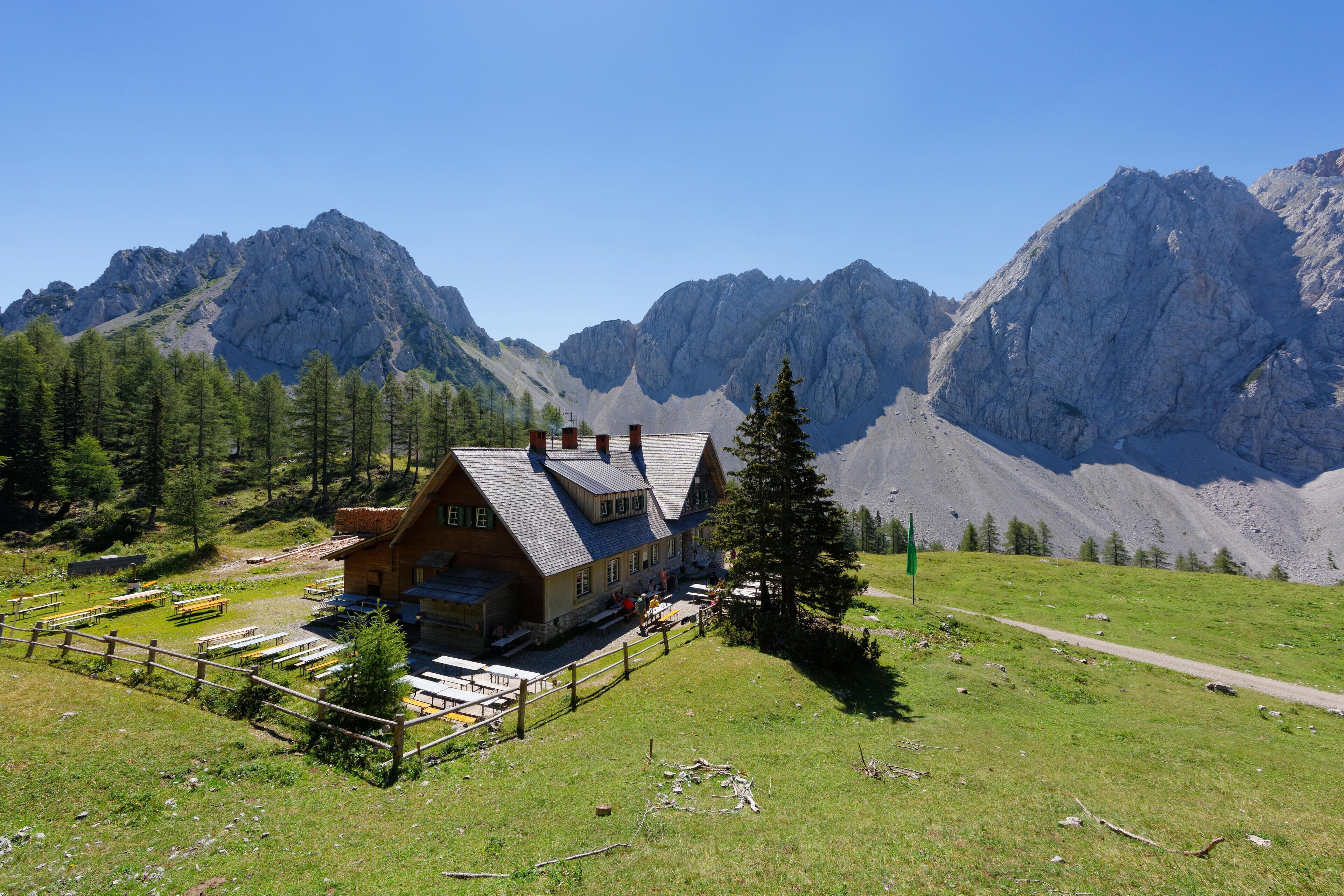 A mountain lodge with a wooden exterior, surrounded by lush greenery and towering rocky mountains.