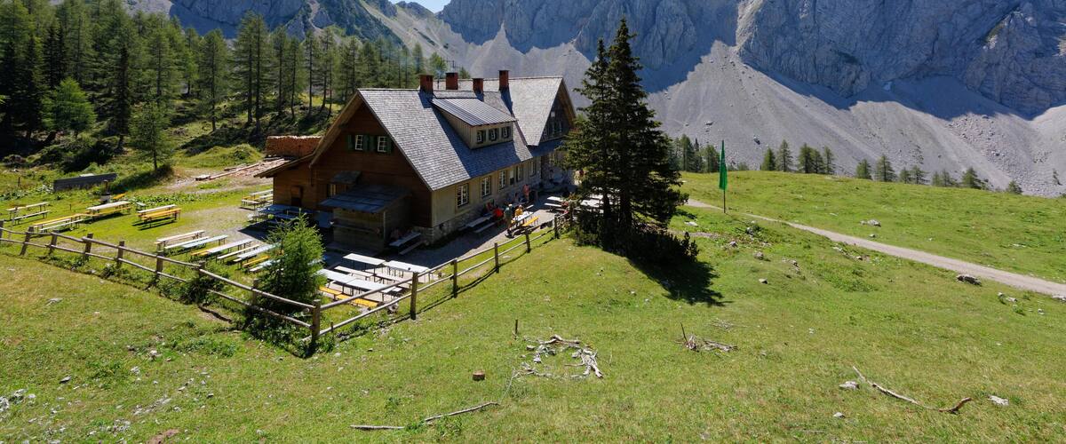 A mountain lodge with a wooden exterior, surrounded by lush greenery and towering rocky mountains.