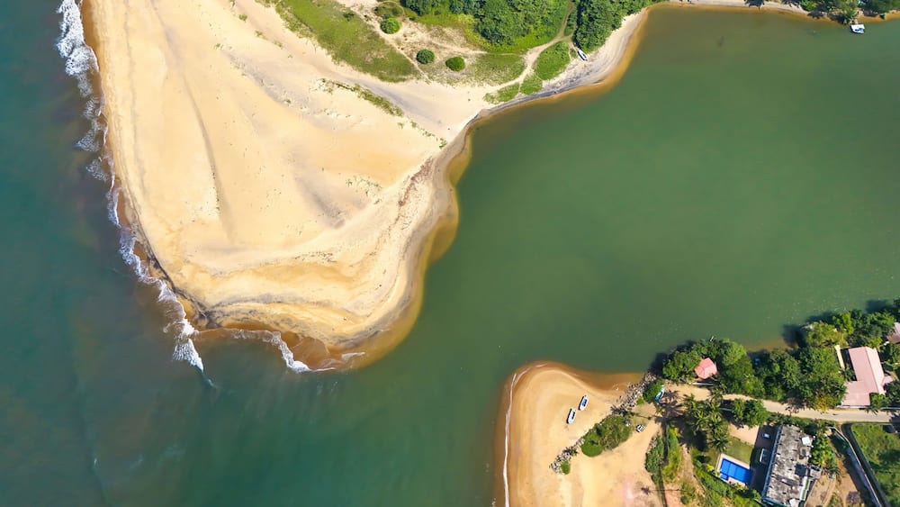 An aerial view of a coastal area with a sandy beach, a green forested area, and a body of water.