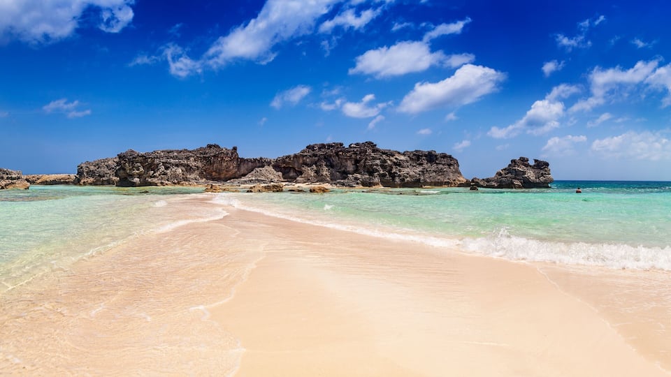 A beach with clear turquoise water, a sandy shore, and rocky formations in the distance.