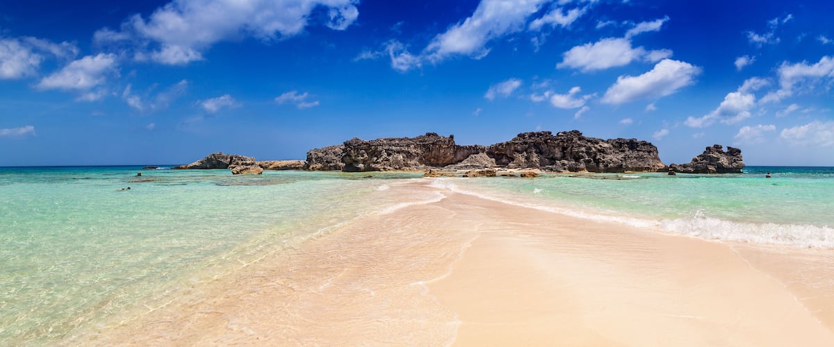 A beach with clear turquoise water, a sandy shore, and rocky formations in the distance.