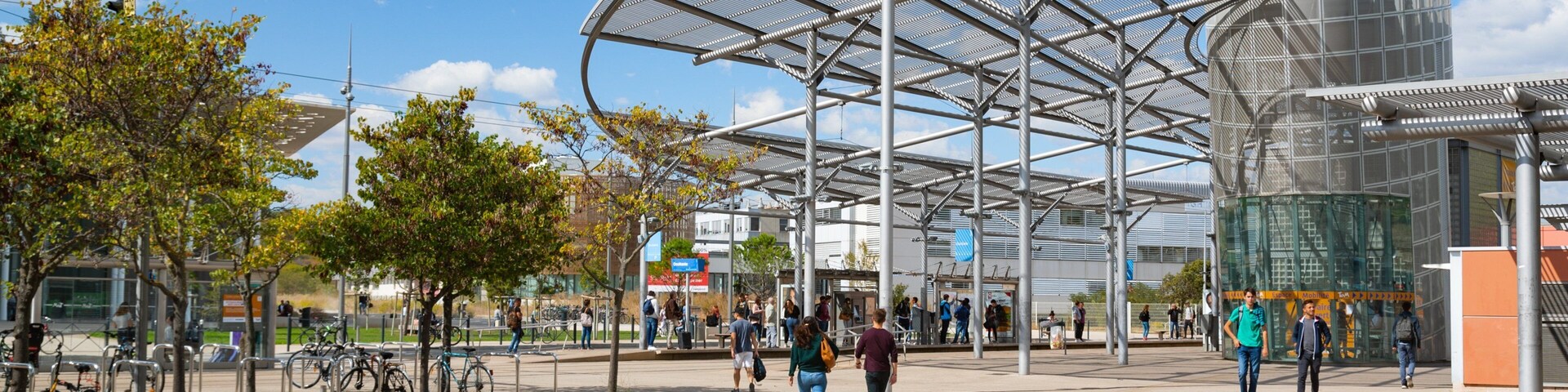 Uma praça pública moderna com um grande toldo metálico, uma fonte e pessoas caminhando.