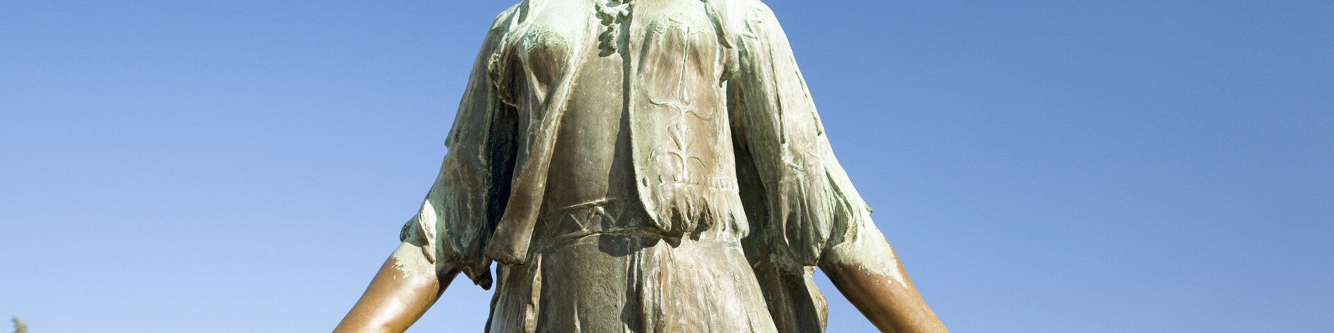 Una estatua con los brazos extendidos, un tocado emplumado y un collar, sobre un cielo azul claro.