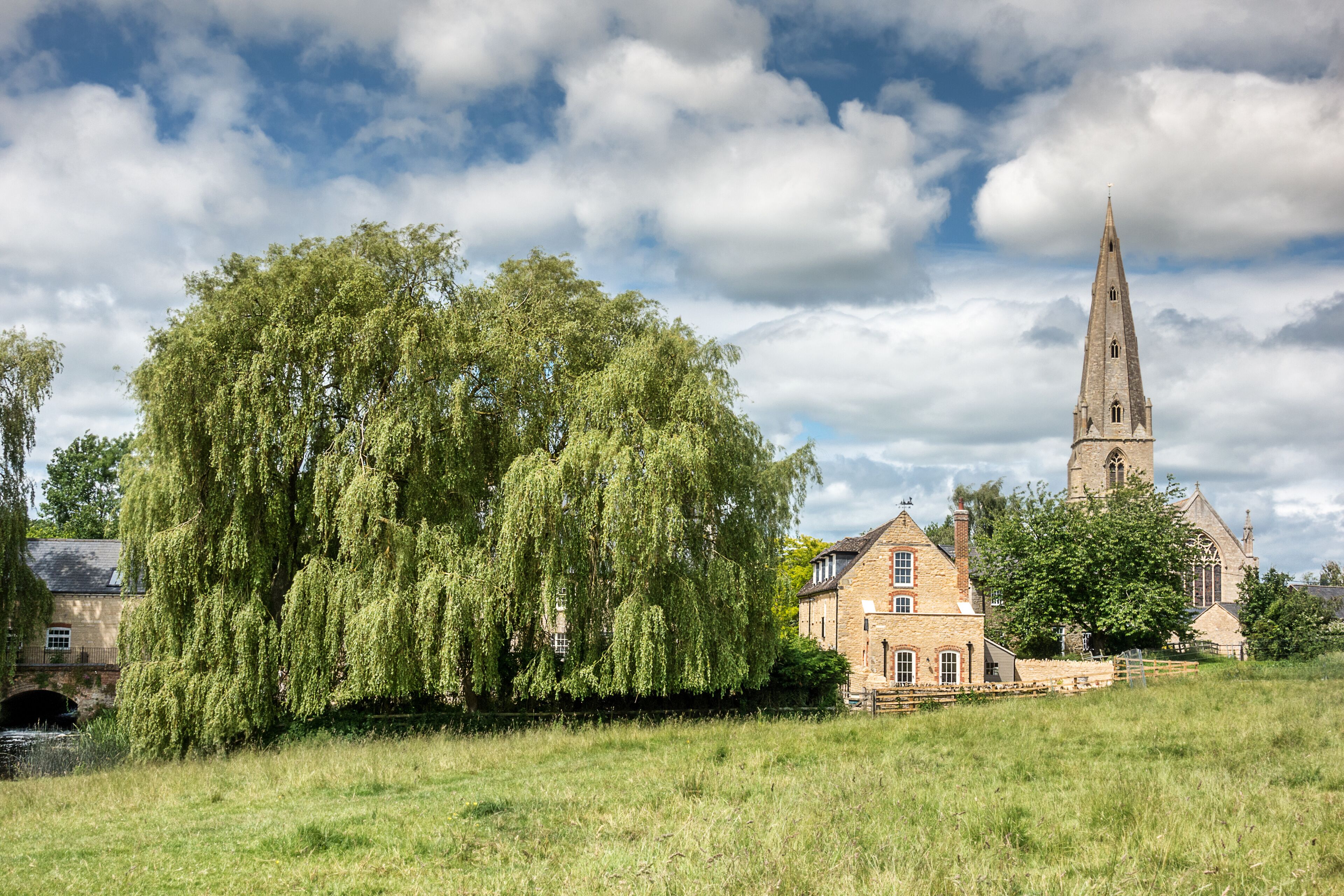 A stone church with a tall spire, a weeping willow tree, and a stone bridge.