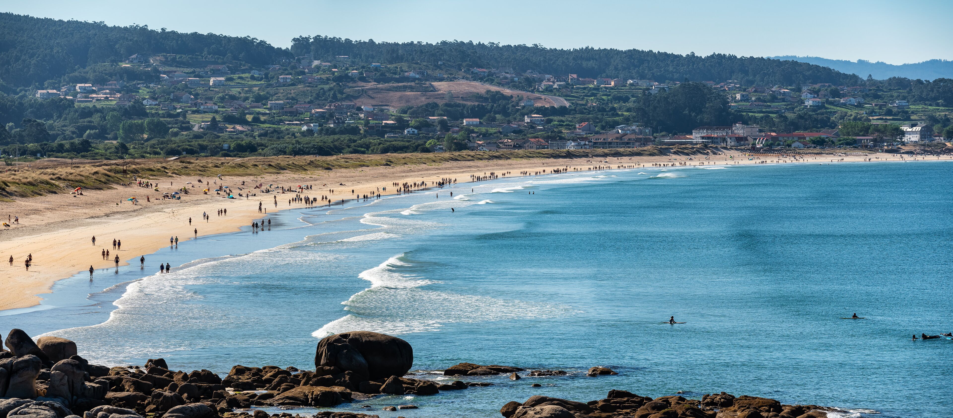 A coastal beach with people, waves, and a town in the background.
