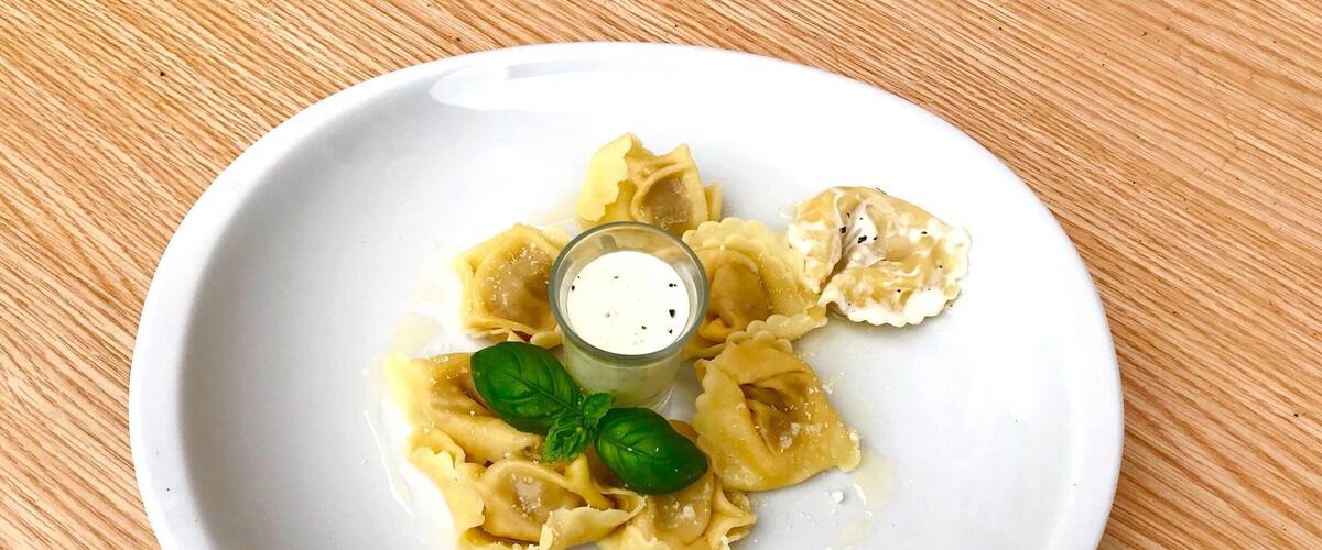 A plate of ravioli with a small bowl of sauce and basil leaves.