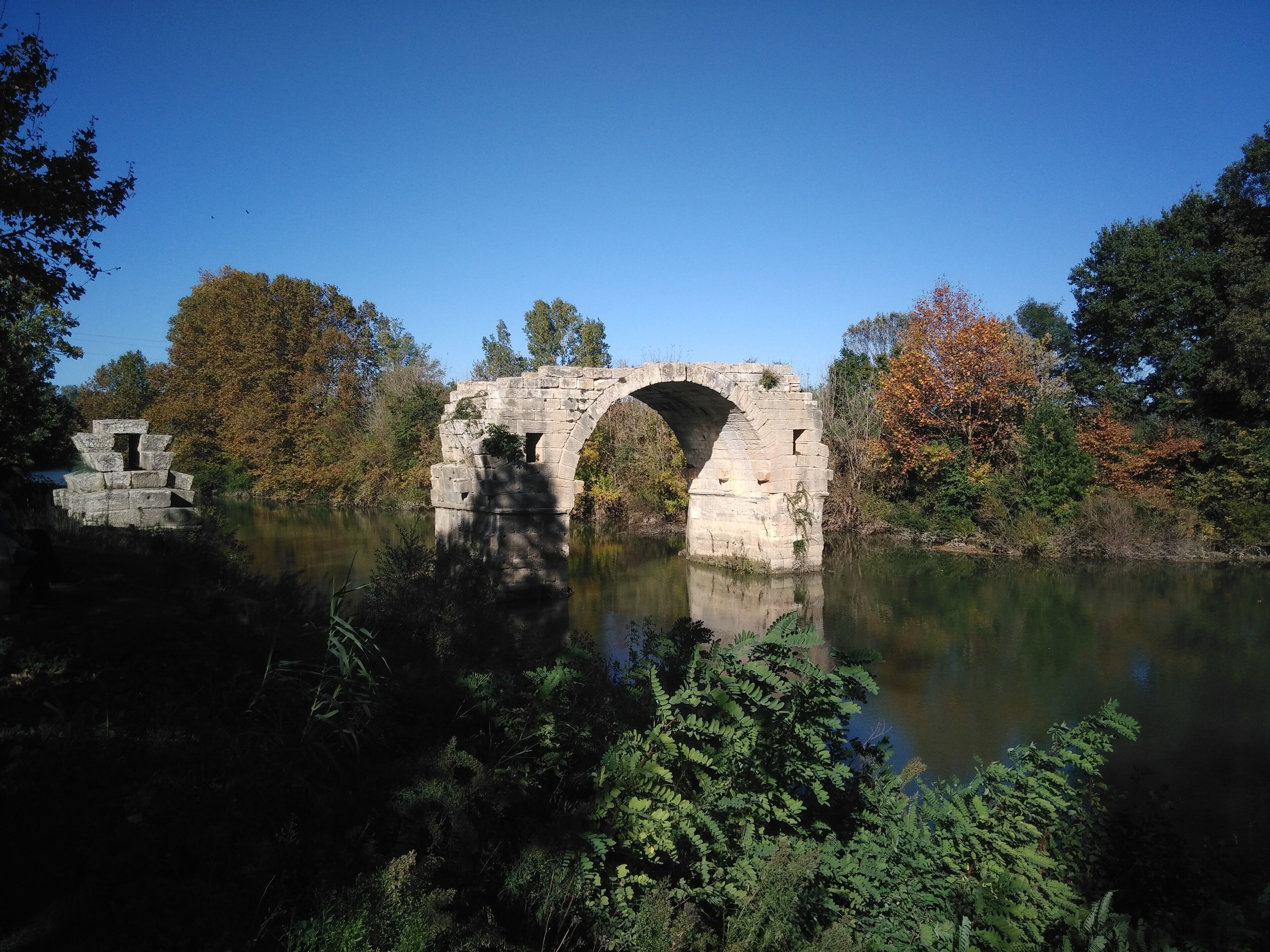 A stone bridge over a calm river, surrounded by autumn foliage and dense greenery.