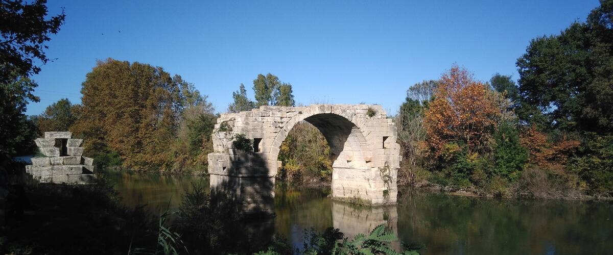 A stone bridge over a calm river, surrounded by autumn foliage and dense greenery.