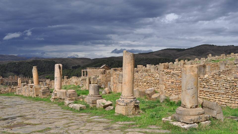 Ancient ruins with stone columns and walls, set against a backdrop of hills and a cloudy sky.