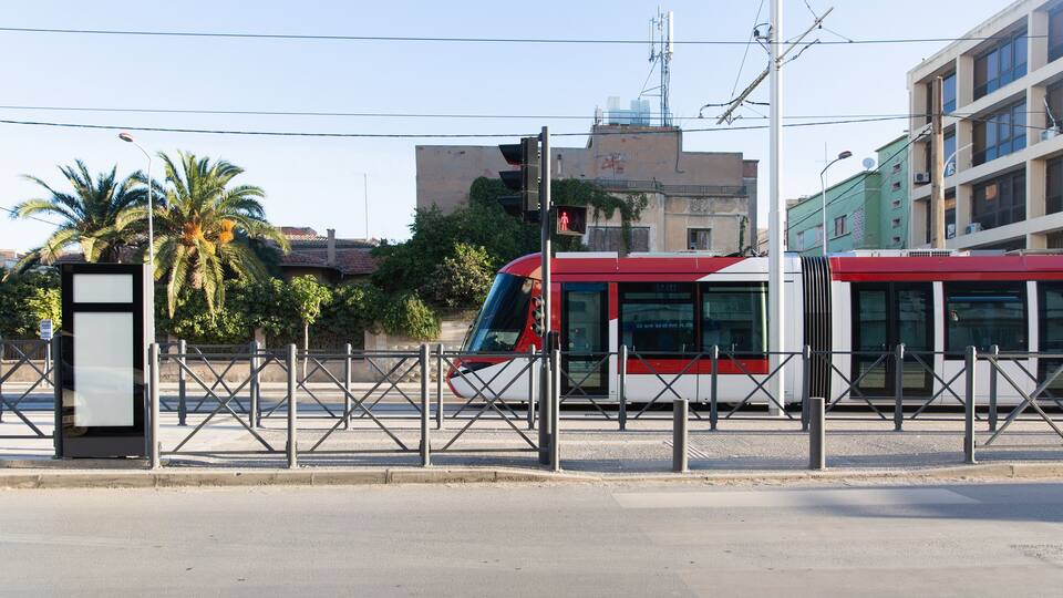 A red and white tram at a station with a fence, palm trees, and buildings in the background.
