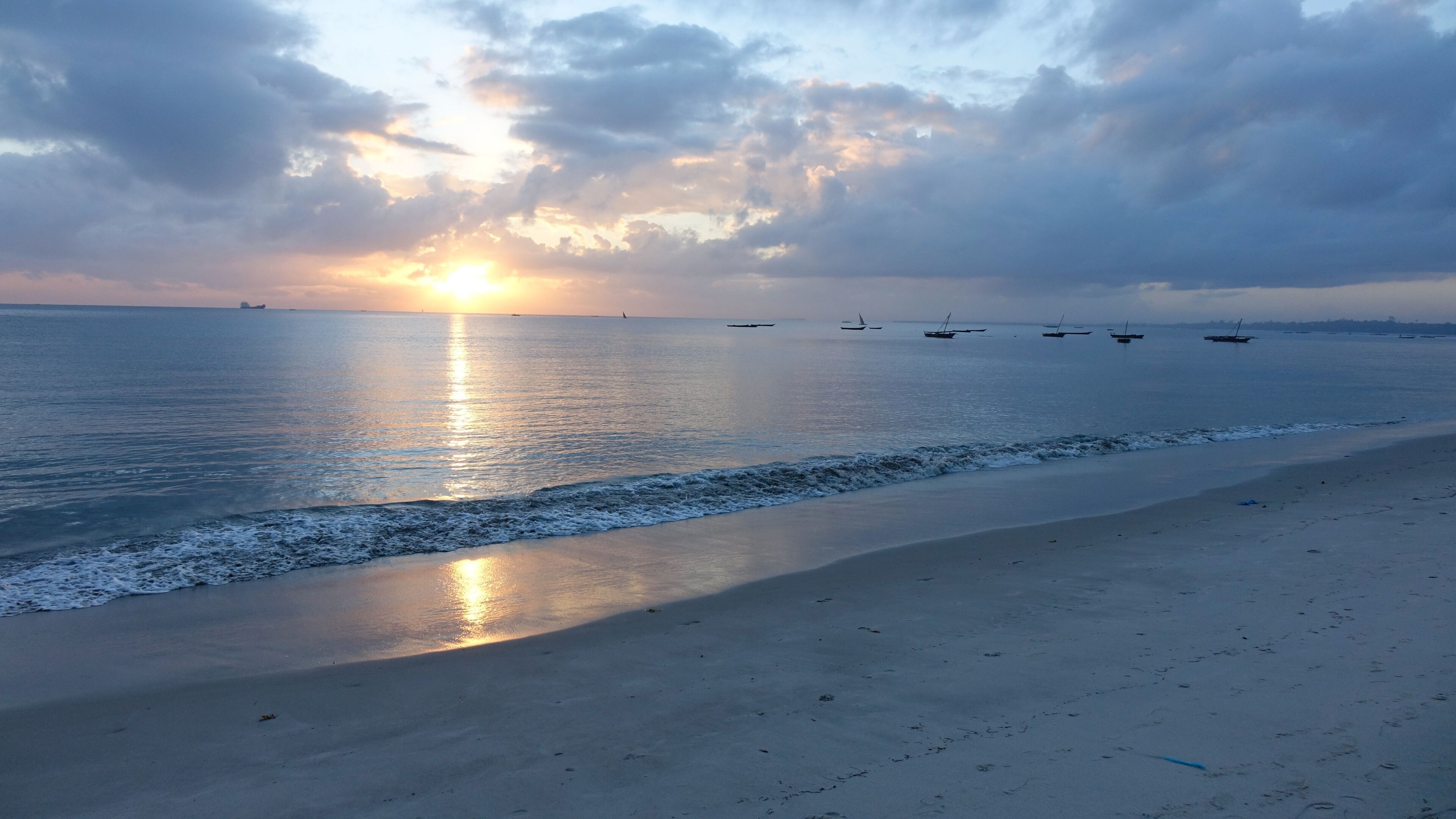 A beach with calm waves, a sunset, and several boats in the distance.