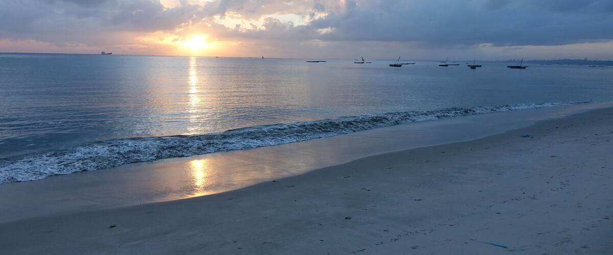A beach with calm waves, a sunset, and several boats in the distance.