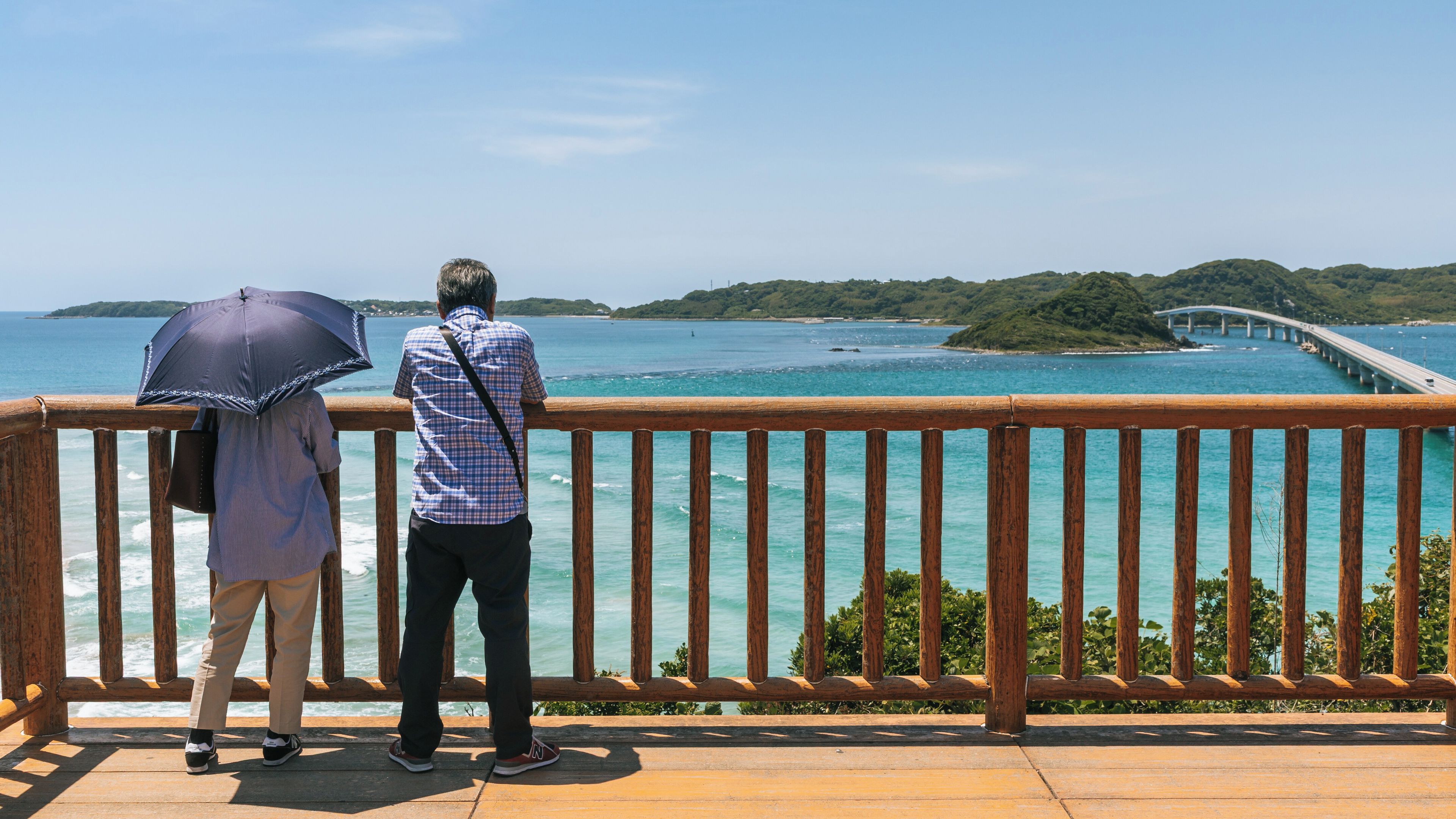 A wooden deck with a railing overlooking turquoise waters, a distant bridge, and green hills.