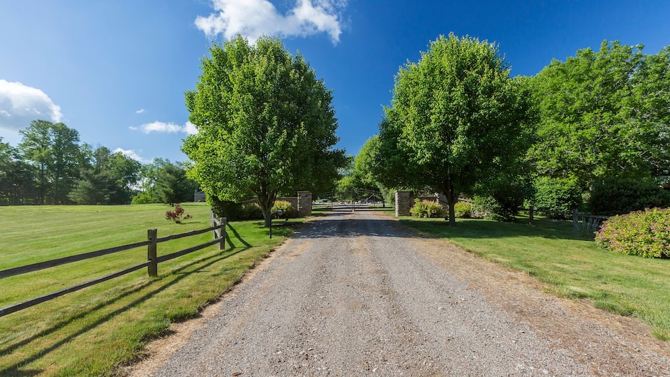 A gravel road flanked by trees and a wooden fence leading to a gated entrance.