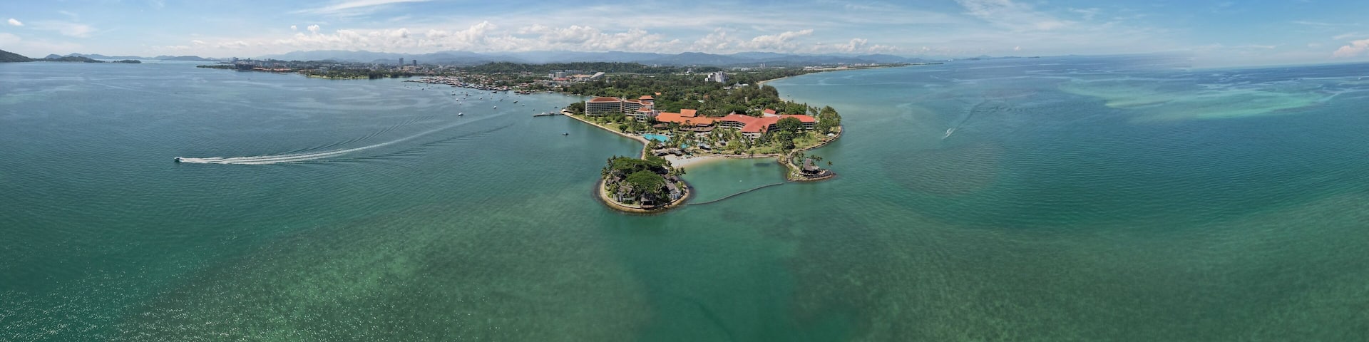 An aerial view of a tropical island with clear turquoise waters, surrounded by a cityscape and distant mountains.
