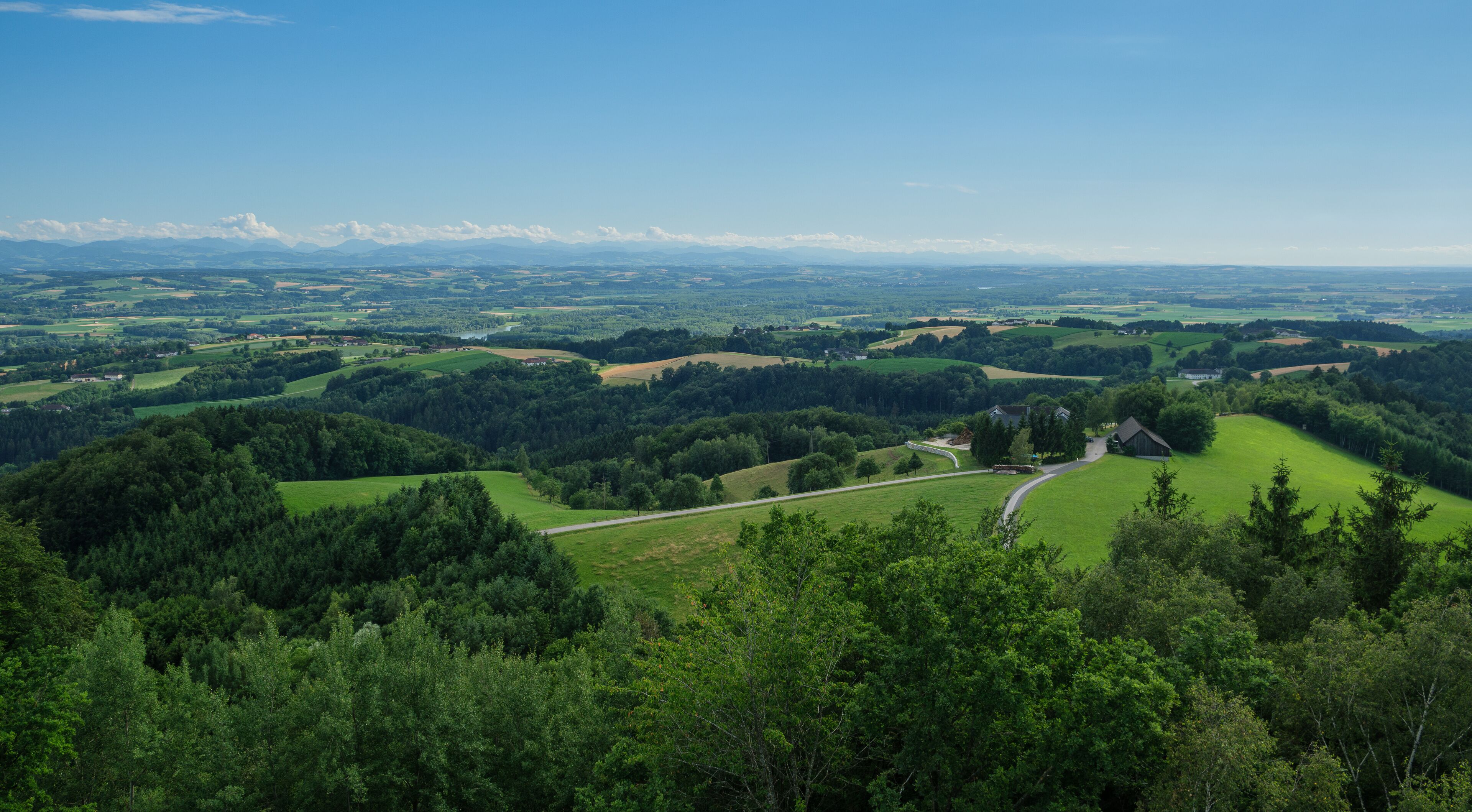 A lush green landscape with rolling hills, a winding road, and a distant mountain range under a clear blue sky.