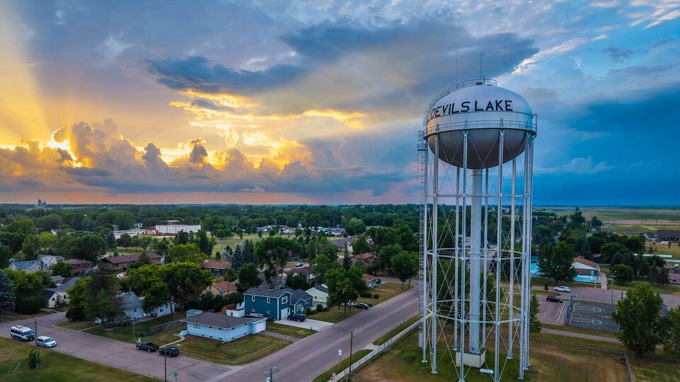 A water tower labeled "DeVilsLake" in a suburban area with houses, roads, and a cloudy sky.