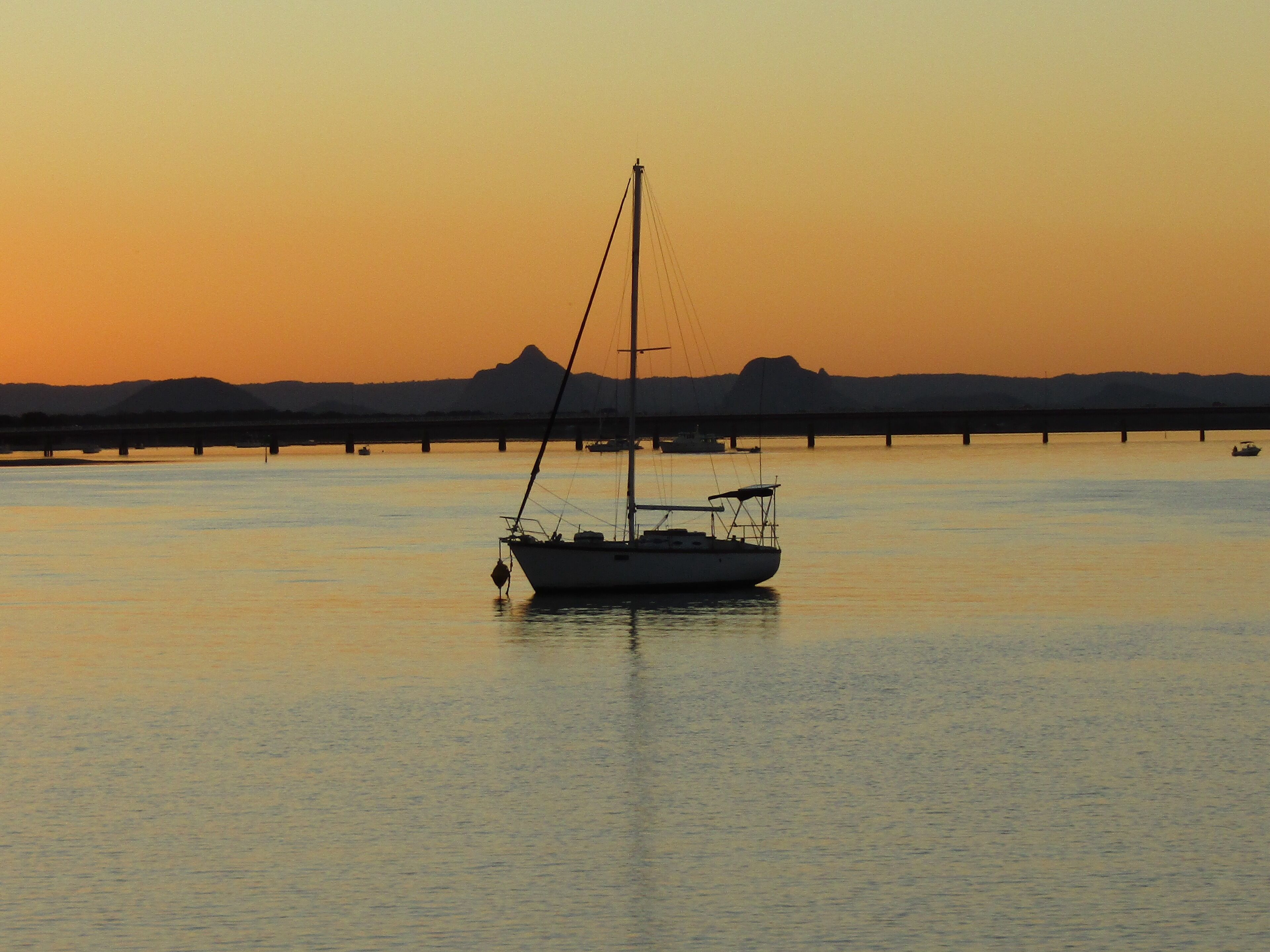 A sailboat anchored in calm waters at sunset with a bridge and distant hills in the background.