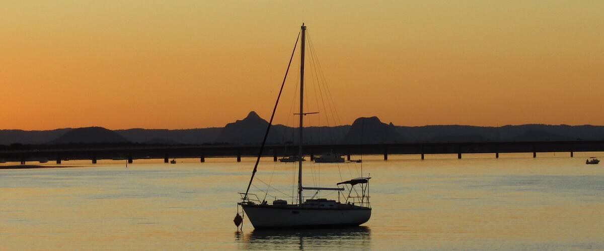 A sailboat anchored in calm waters at sunset with a bridge and distant hills in the background.