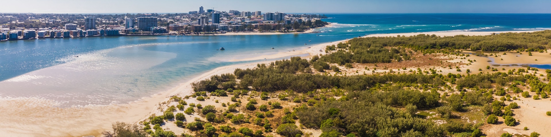 A coastal cityscape with a beach, dense greenery, and a clear blue sky.