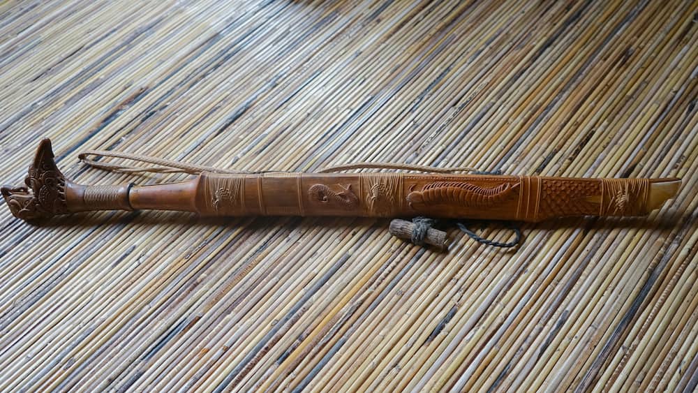 A wooden walking stick with intricate carvings and a rope handle, lying on a bamboo mat.