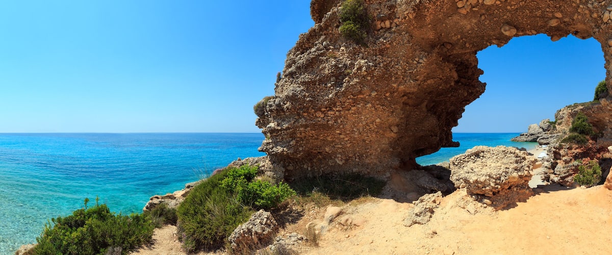 A coastal landscape with a clear blue sea, a sandy beach, and a rocky cliff with a natural arch formation.