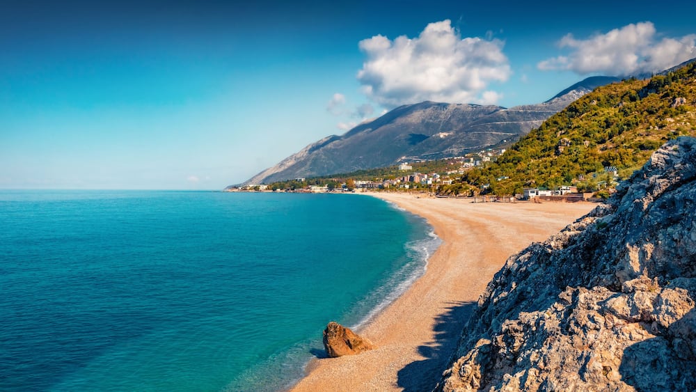 Un paysage côtier avec une plage de sable, des eaux turquoise et limpides, et un arrière-plan montagneux.