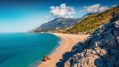 Paisagem costeira com praia arenosa, águas turquesa cristalinas e montanhas ao fundo.