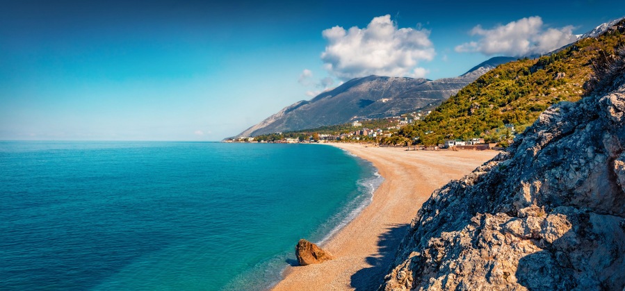 Un paysage côtier avec une plage de sable, des eaux turquoise et limpides, et un arrière-plan montagneux.