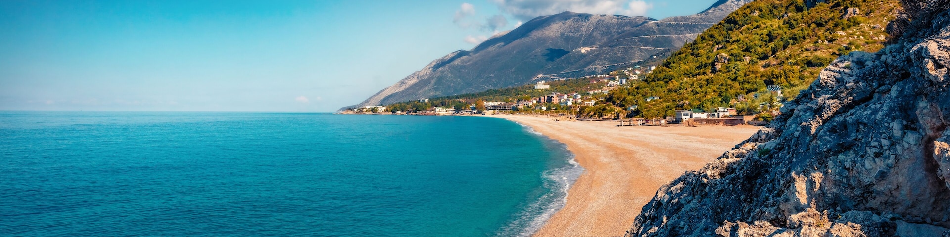A coastal landscape with a sandy beach, clear turquoise waters, and a mountainous backdrop.