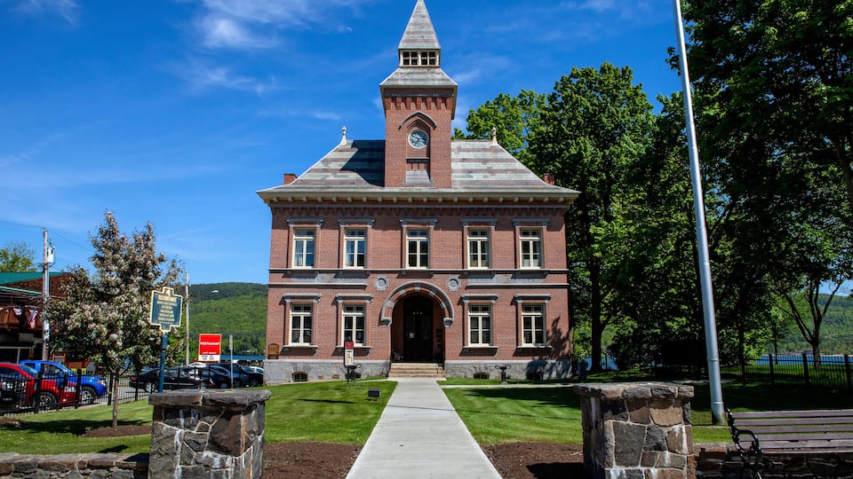 A historic brick building with a clock tower, a paved walkway, and a park with a bench.