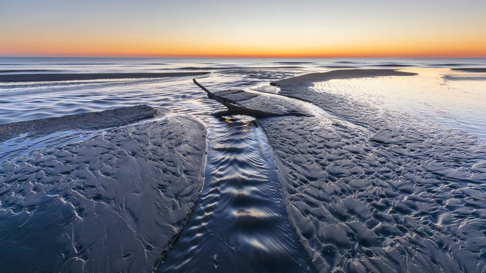 A beach at sunset with calm waves and a clear sky.