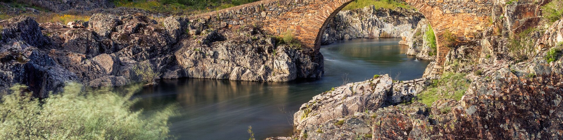 Un puente de piedra sobre un río tranquilo, rodeado de terreno rocoso y vegetación.