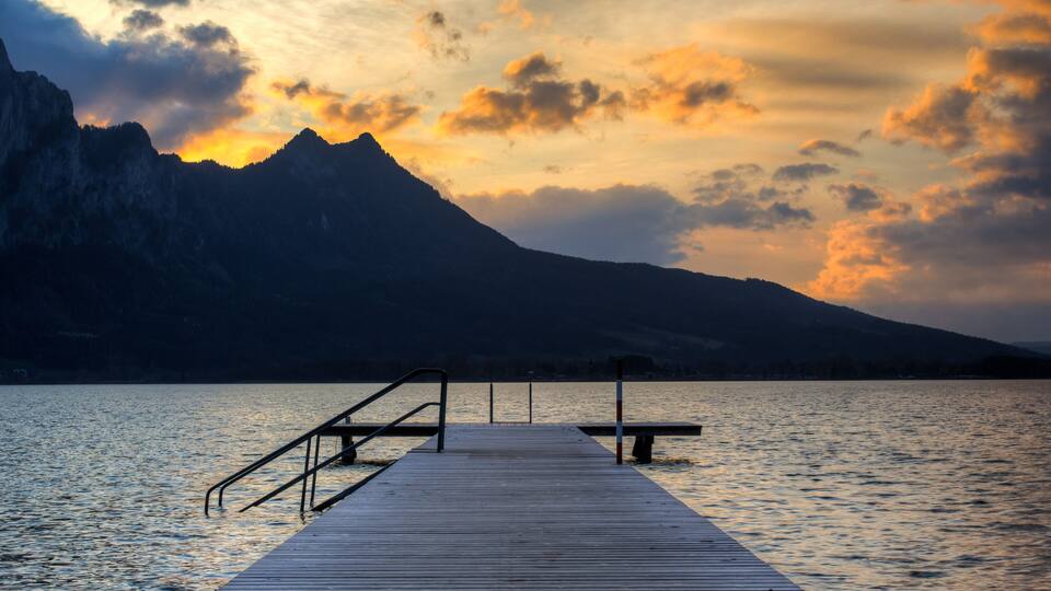 A wooden pier extending into calm water, with a mountain in the background and a sunset sky.