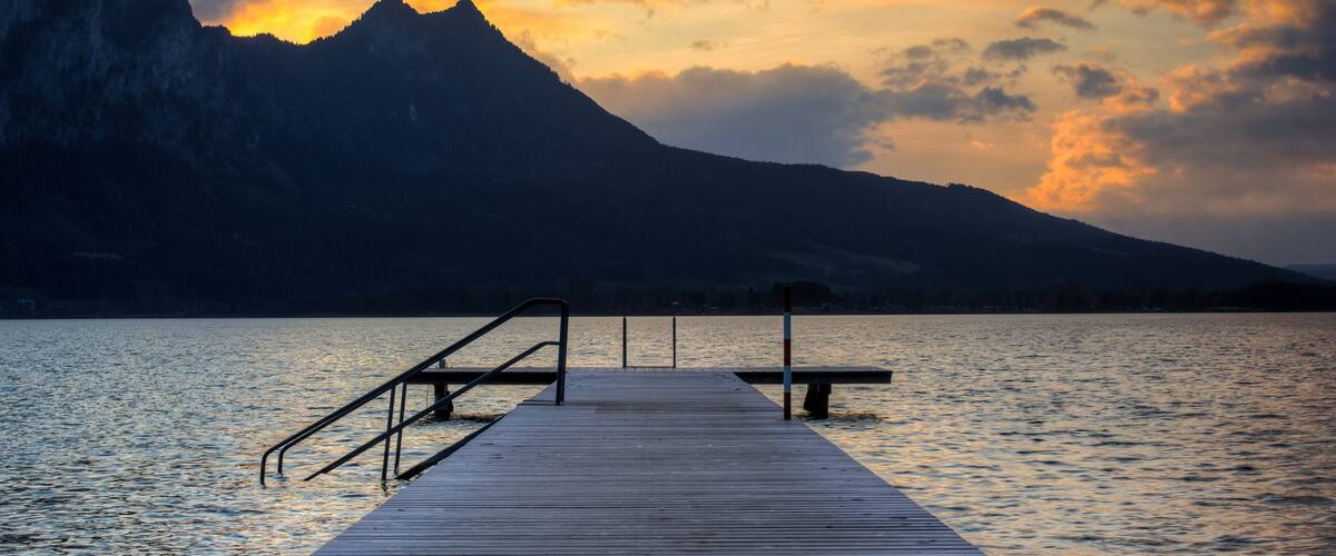 A wooden pier extending into calm water, with a mountain in the background and a sunset sky.