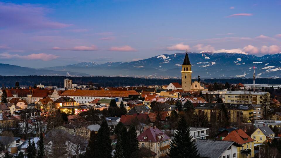 A town with a prominent church tower, snow-capped mountains in the background, and a mix of traditional and modern buildings.