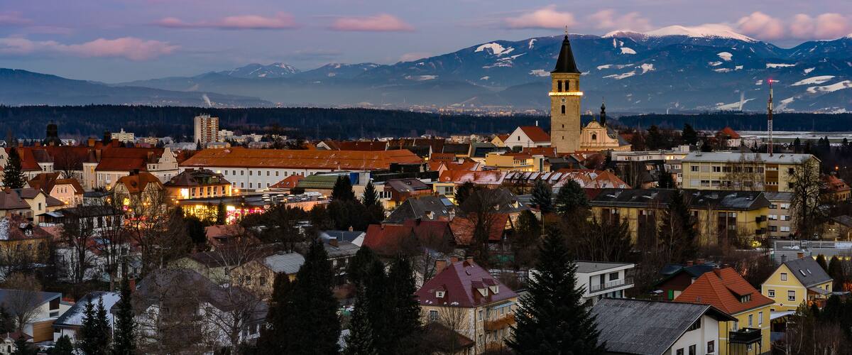 A town with a prominent church tower, snow-capped mountains in the background, and a mix of traditional and modern buildings.