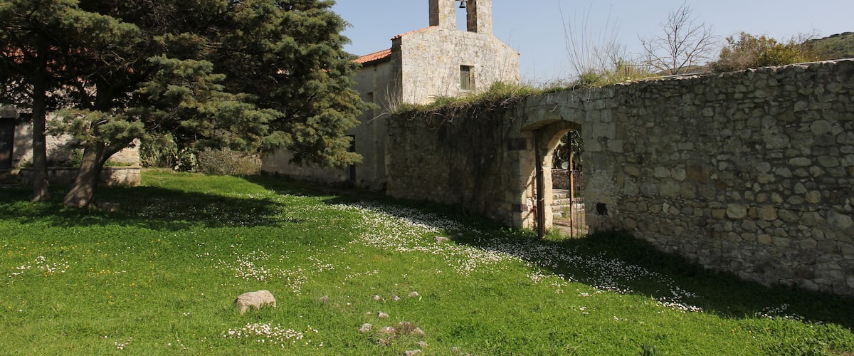 An old stone church with a bell tower, surrounded by a stone wall and a grassy area with scattered white flowers.