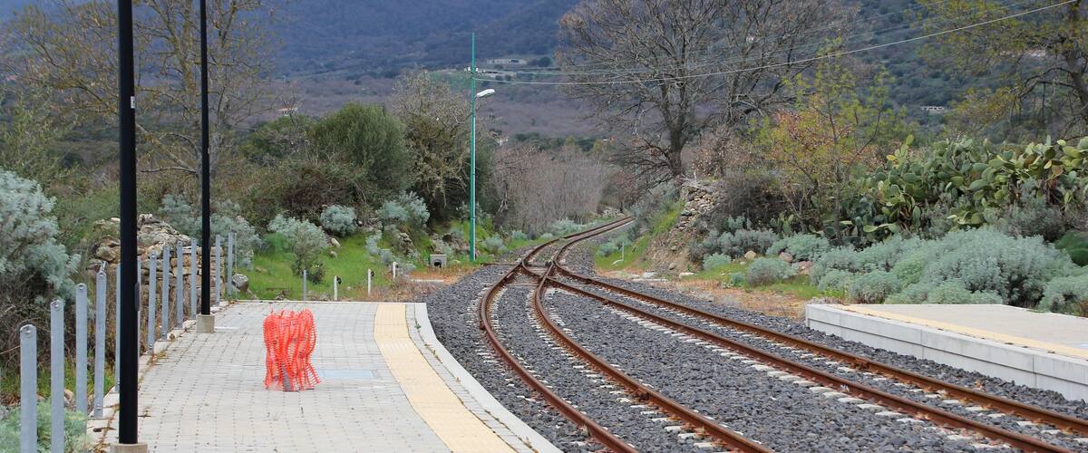 A railway track with a pedestrian walkway, surrounded by greenery and hills in the background.
