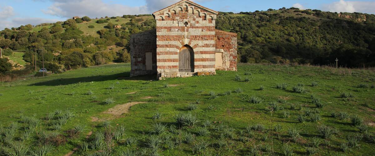 A small brick structure with a triangular roof and arched doorway, surrounded by a grassy field and hills in the background.
