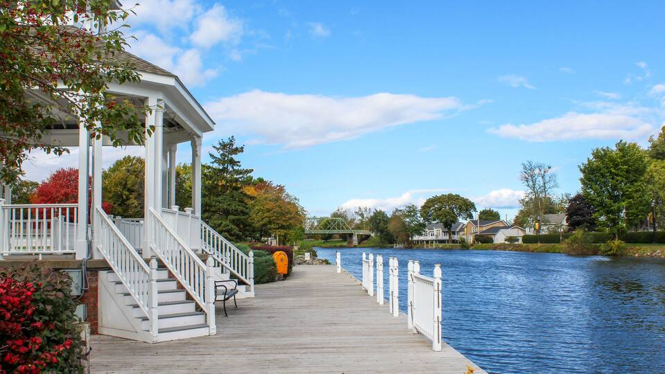 A wooden pier extending over a calm river, with a gazebo on the left and houses in the background.