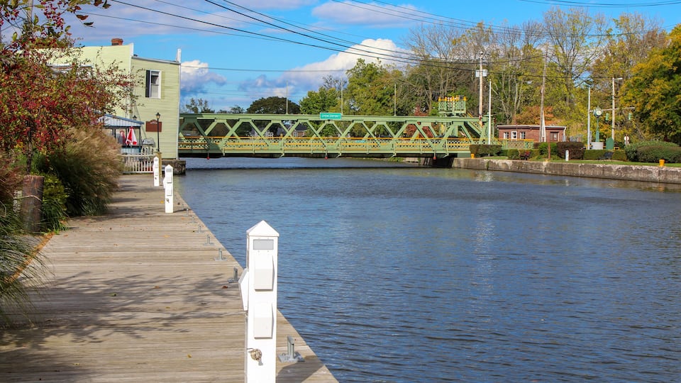 A wooden boardwalk beside a calm river, a green bridge, and a small building with a red roof.
