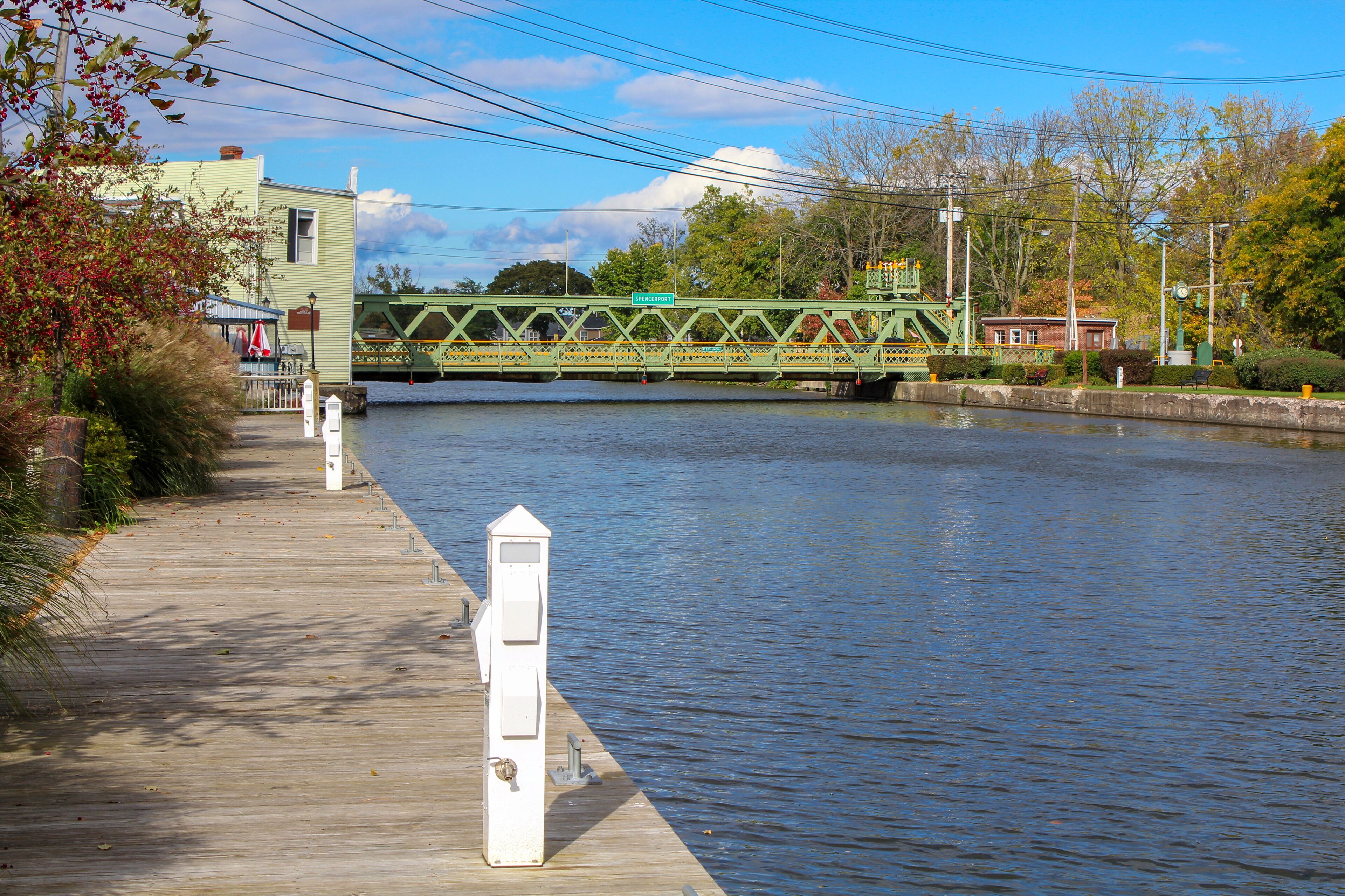A wooden boardwalk beside a calm river, a green bridge, and a small building with a red roof.