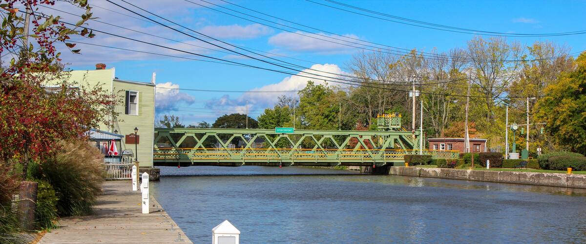 A wooden boardwalk beside a calm river, a green bridge, and a small building with a red roof.