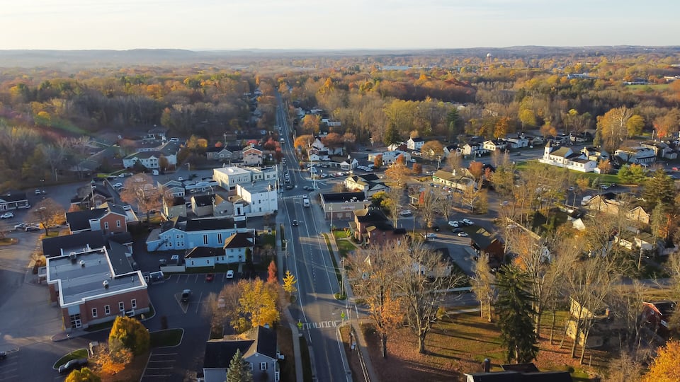 An aerial view of a town with a mix of residential and commercial buildings, surrounded by autumn-colored trees.
