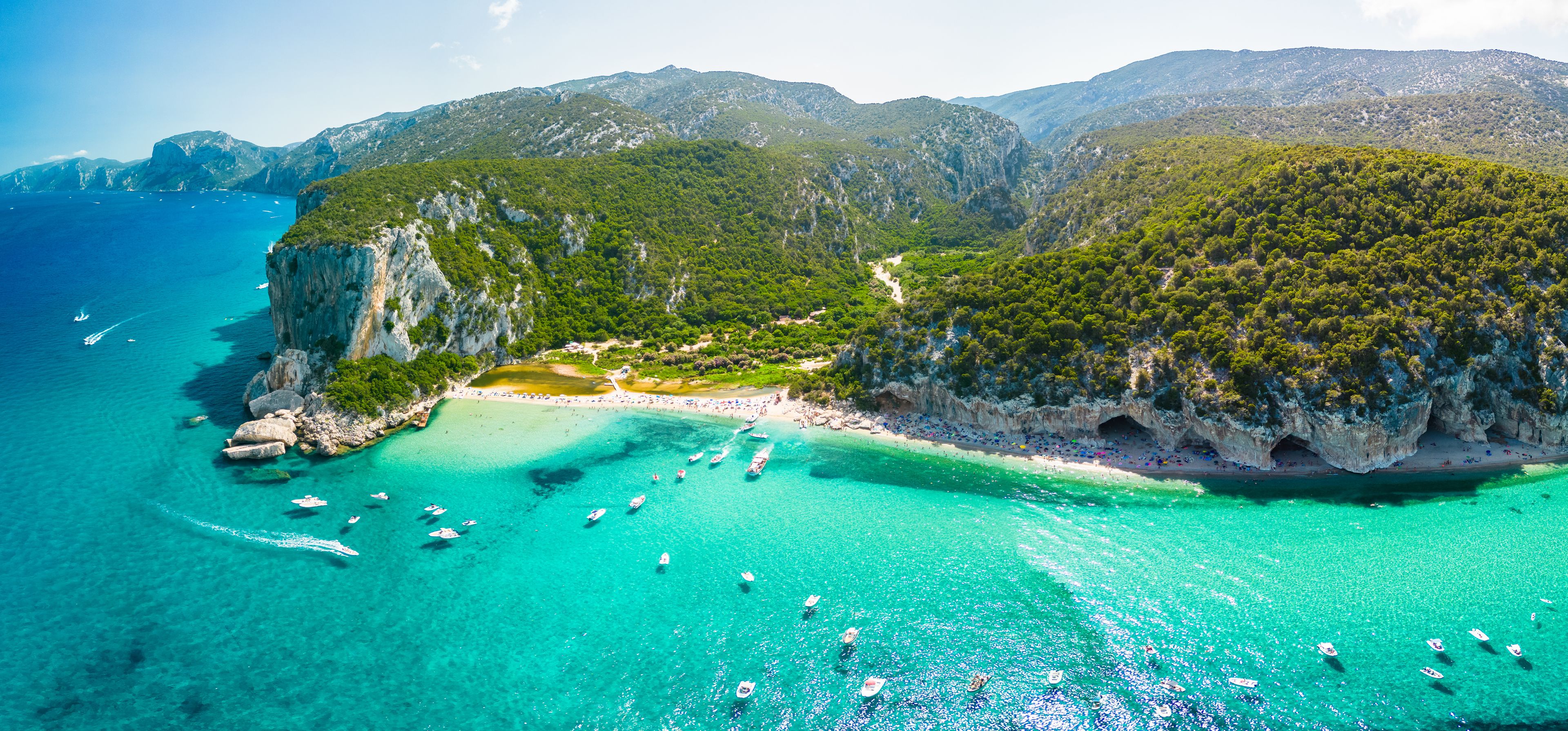 A coastal landscape with clear turquoise waters, a sandy beach, and lush green hills.